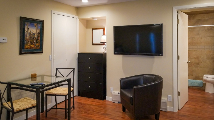 Interior view of the cozy guest room in HA Whitney Inn, featuring a dining table with chairs, a dresser, a TV mounted on the wall, and a private bathroom.